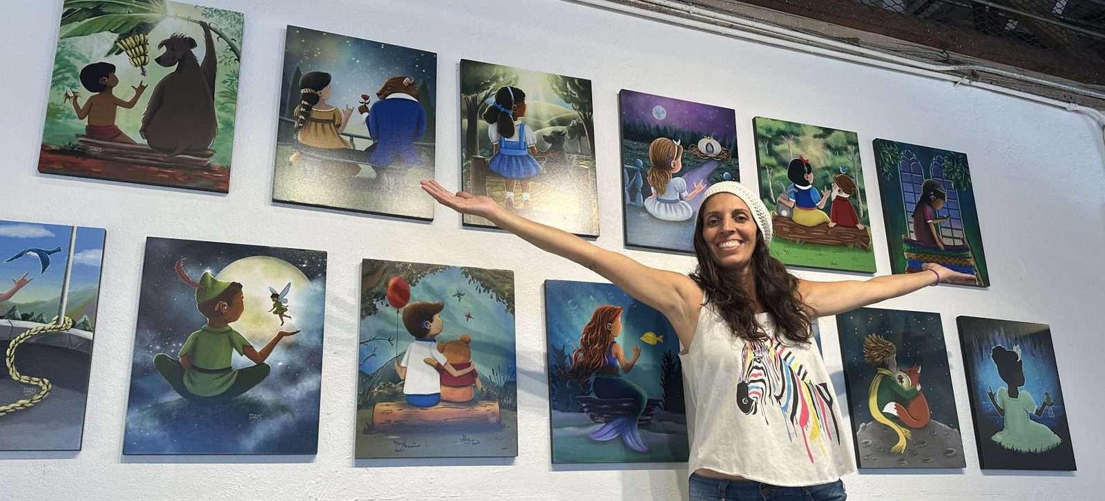 Artist Priscila Soares in front of her collection of Fairy Tale art with characters wearing cochlear implants, hearing aids, bone anchored hearing and sign language